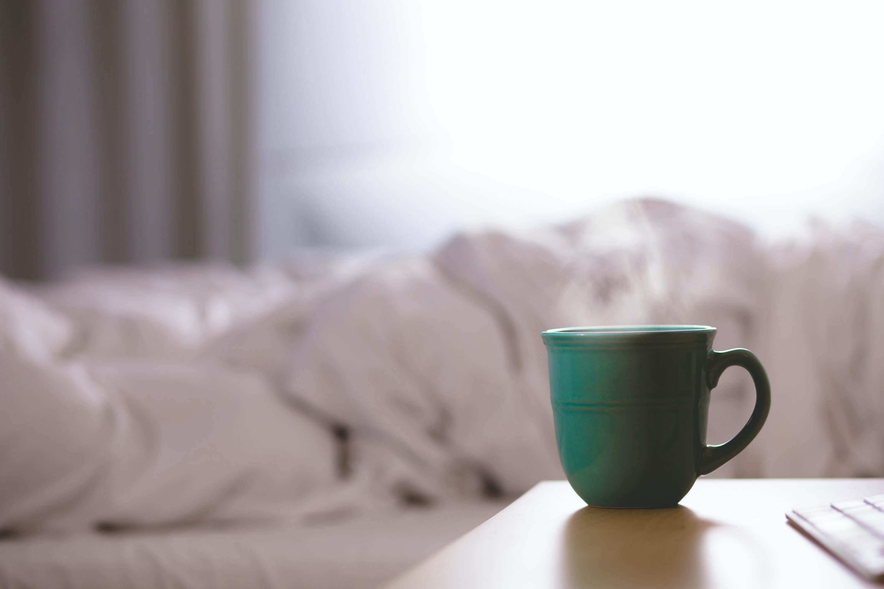 A cup of hot coffee on a bed side table with a slept in bed in the background