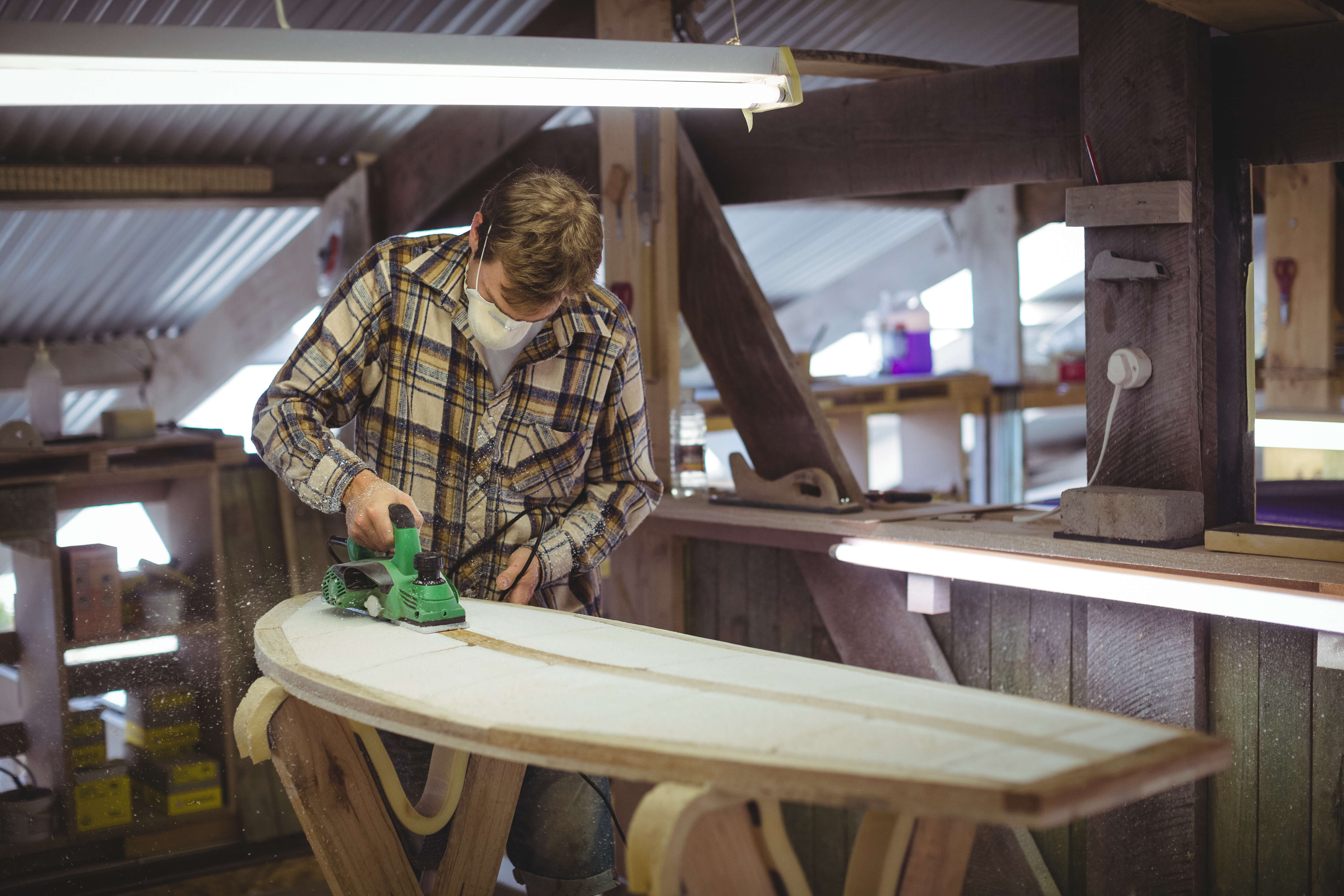 Full length photo of a man using a powertool to work on a surfboard in a workshop