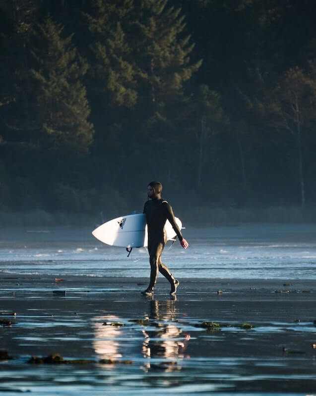 A surfer in a hooded wetsuit walking out of the ocean carrying a surfboard.