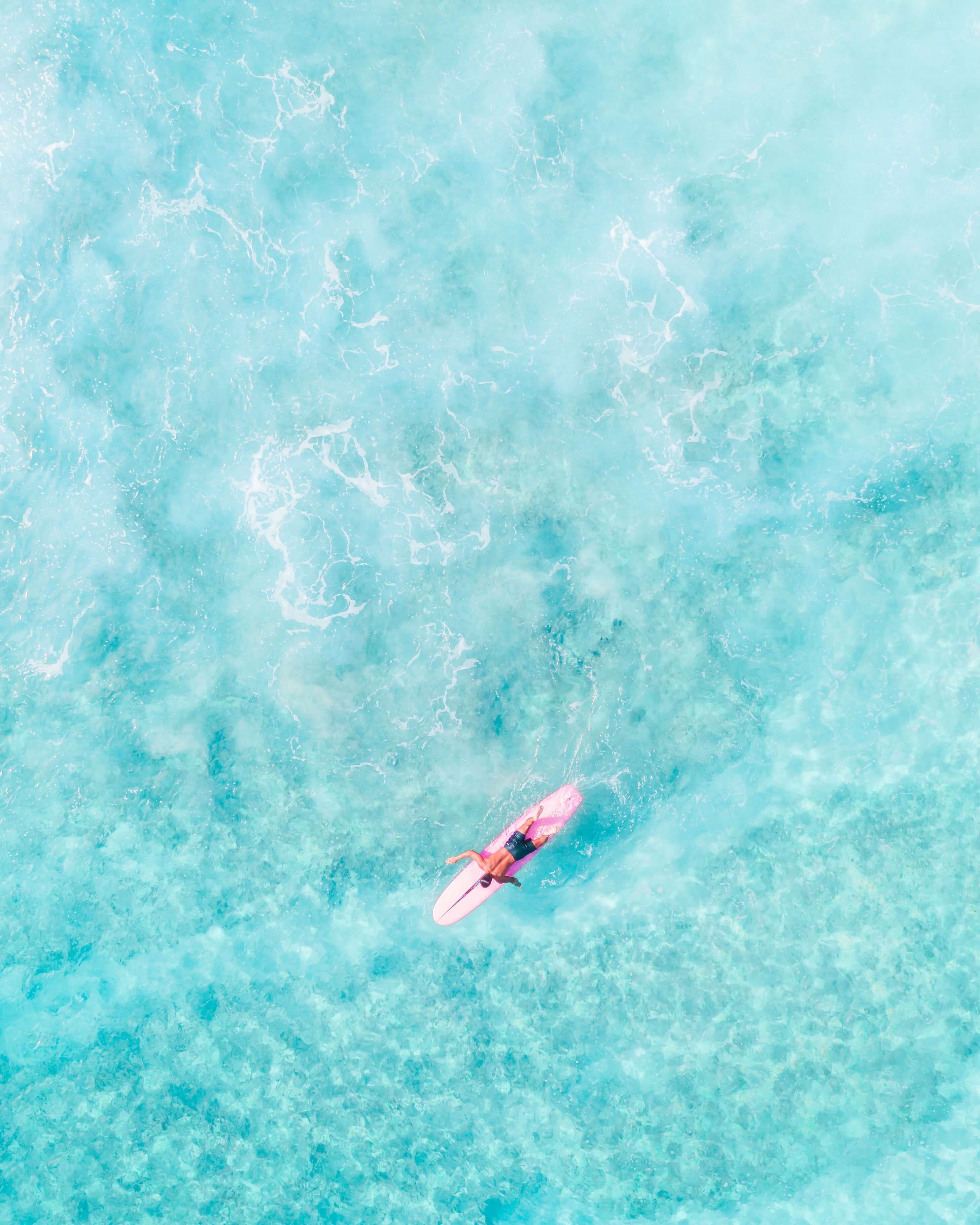 Birds eye view of a surfer with a pink longboard paddling in clear blue water 