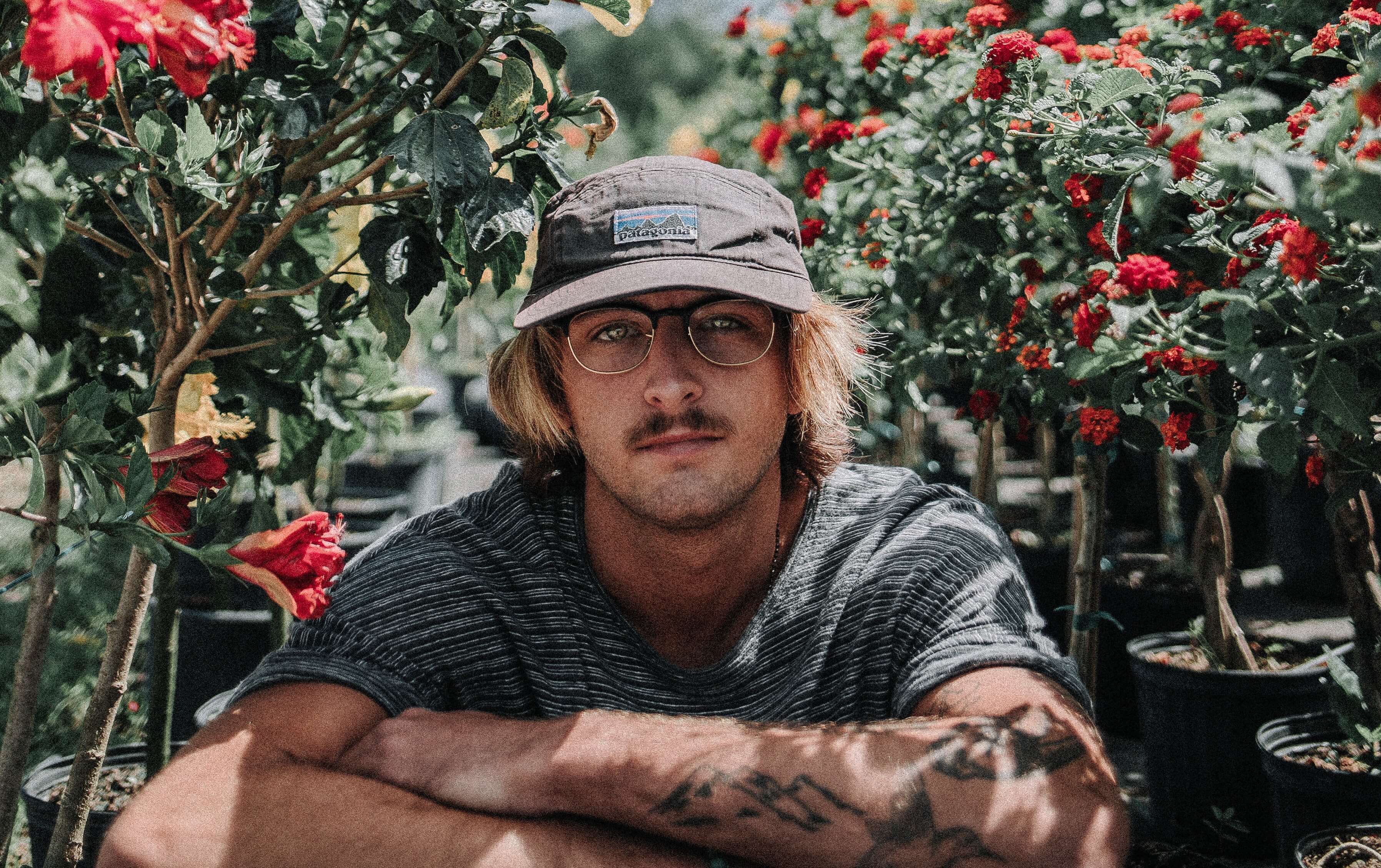 Head shot of a man with tattoos wearing a hat and glasses sat in some shrubbery